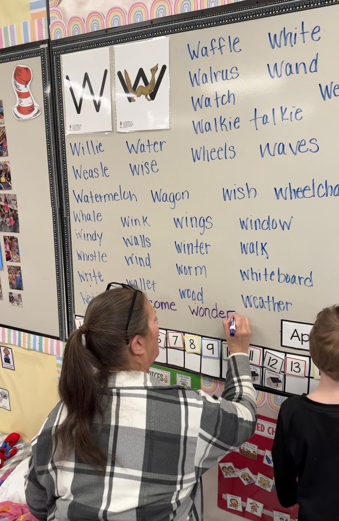 A preschool teacher writes additional “W” words on a classroom word wall while a child stands nearby observing and participating.