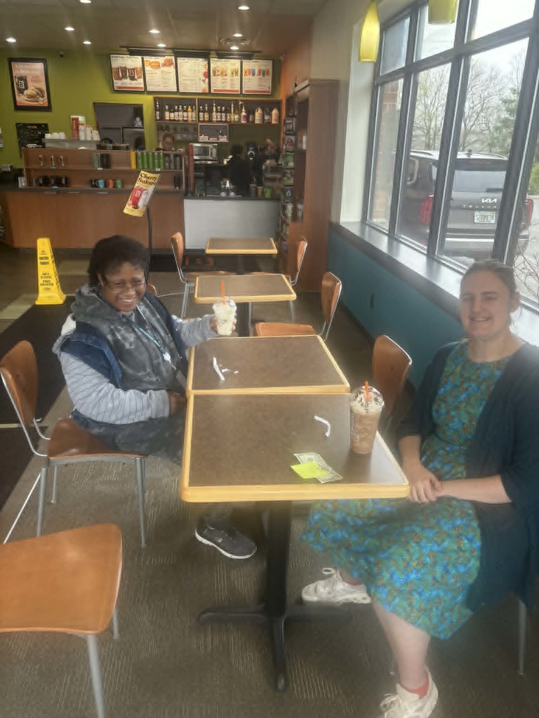 Two students sit together at Biggby Coffee, smiling and holding their drinks as they chat and build friendship skills.