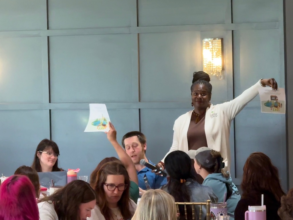 An educator reads a Tucker the Turtle story into a microphone while others hold up related visuals during a professional development activity.