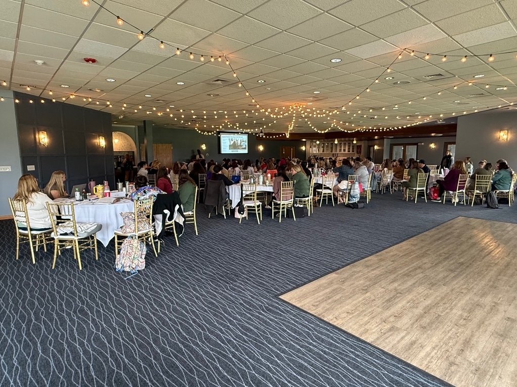 A large group of early childhood educators sit at round tables in a conference room with string lights, participating in a professional development session.