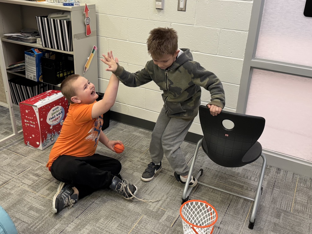 Two young boys celebrating a successful shot with a high five in a classroom. One boy, wearing an orange shirt, sits on the carpeted floor holding a small toy basketball; the other boy, in a camouflage hoodie, stands next to him, leaning on a black chair. A small white and orange toy basketball hoop sits on the floor in front of them.Two young boys celebrating a successful shot with a high five in a classroom. One boy, wearing an orange shirt, sits on the carpeted floor holding a small toy basketball; the other boy, in a camouflage hoodie, stands next to him, leaning on a black chair. A small white and orange toy basketball hoop sits on the floor in front of them.