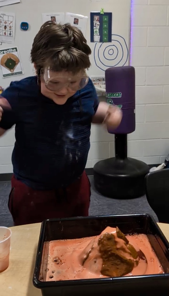 A young boy in a blue shirt and safety goggles reacts with excitement and a wide smile as a model volcano erupts in front of him. He is jumping or moving his arms with joy while looking down at the black tray filled with bubbling orange foam. In the background of the classroom,  educational posters are visible against a white brick wall.