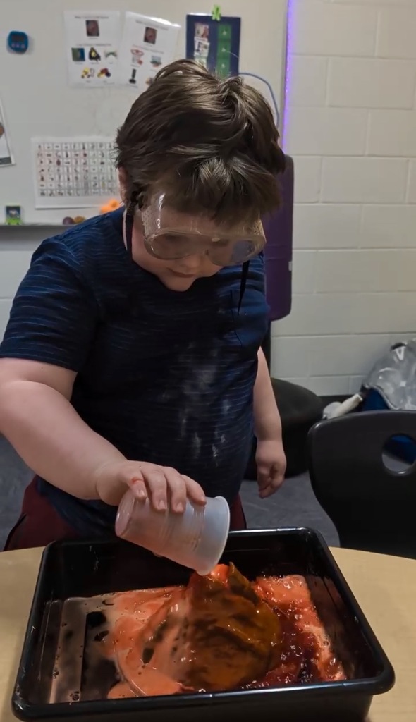 A young boy wearing large clear safety goggles focuses intently as he pours liquid from a small plastic cup onto a model volcano. The volcano is erupting with bubbly, orange-red foam that spills into a black rectangular tray. He is wearing a blue textured t-shirt, and the background shows a classroom setting with a white brick wall and educational posters.