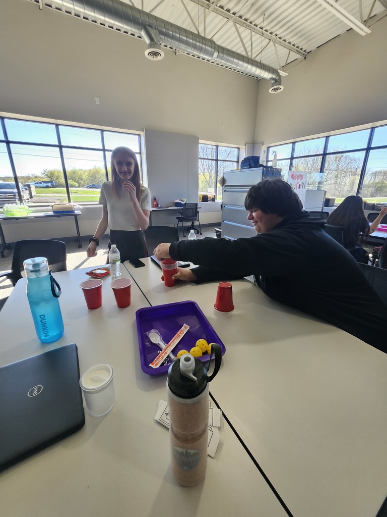 Two students sit at a white table in a bright, modern classroom or office space, engaged in an activity with red plastic cups and small yellow balls on a purple tray. The room features large windows showing a parking lot and greenery, with exposed ductwork on the ceiling.