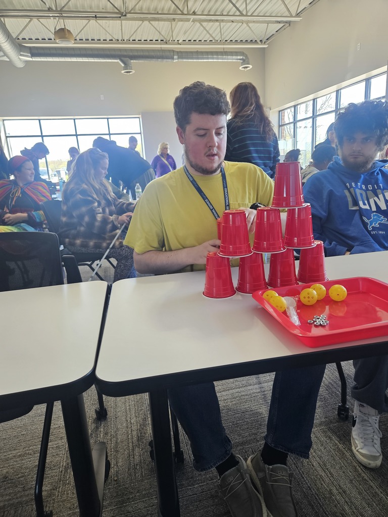 A young man in a yellow t-shirt focuses on stacking red cups into a pyramid on a white table. A red tray with yellow practice golf balls sits nearby.