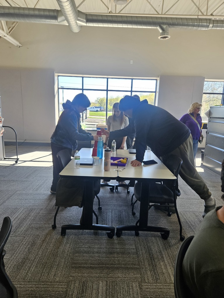 Two students stand at a table and work together to build a tower of red plastic cups in a large, bright room with high ceilings and industrial ductwork.