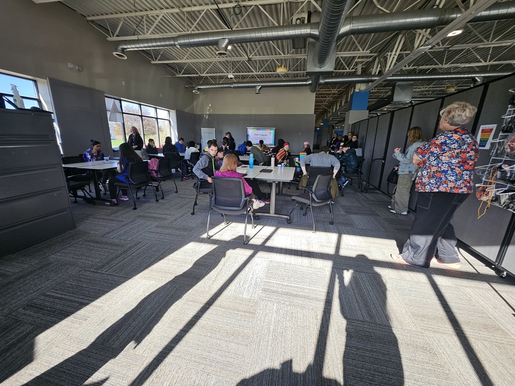 A wide view of a large, modern educational space where groups of students and staff are gathered at various tables for a presentation or workshop. Long shadows stretch across the carpeted floor from the large windows.