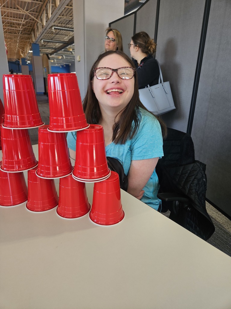 A smiling young woman wearing glasses sits at a table behind a stacked pyramid of red plastic cups. Two other women stand in the background of the bright, open room.