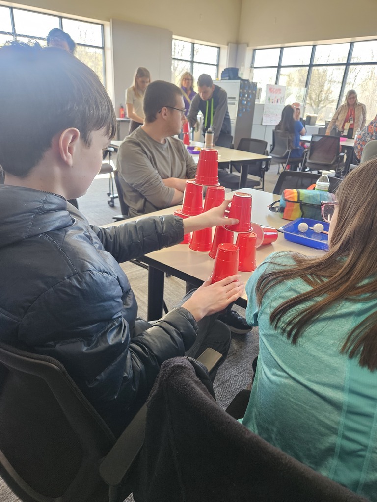 : A student in a black puffy jacket stacks red plastic cups into a pyramid at a table, while other students and staff participate in the activity in a sunlit classroom.