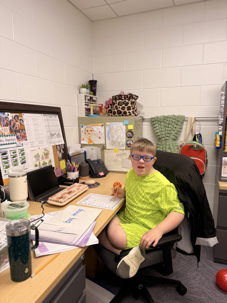 A young boy with Down syndrome, wearing blue-rimmed glasses and a bright neon green outfit, sits at a desk in an office setting, smiling toward the camera while holding a pencil.
