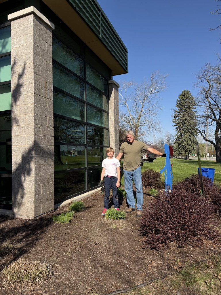 A full-length vertical shot shows an older man with graying hair and a younger boy standing together in a landscaped bed next to a modern building.  The man wears an olive green T-shirt, blue jeans, and work boots. He has his left arm extended, touching a blue, human-shaped metal cutout decoration that stands in the garden bed. The boy stands to his right, wearing a white T-shirt, dark blue pants, and pink and red sneakers. He holds a pair of dark work gloves in his right hand.  The building to their left features a large glass wall with dark metal framing and a tall pillar made of light-colored textured masonry blocks. The ground is covered in dark mulch with several small green plants and a large reddish-purple bush in the foreground. In the background, there is a lush green lawn, several bare and evergreen trees, and a clear blue sky.