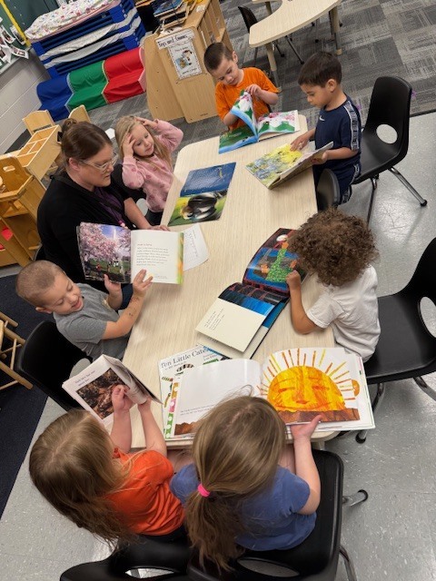 A preschool teacher sits at a table with a group of children, each looking at books together and discussing features like pictures and print.