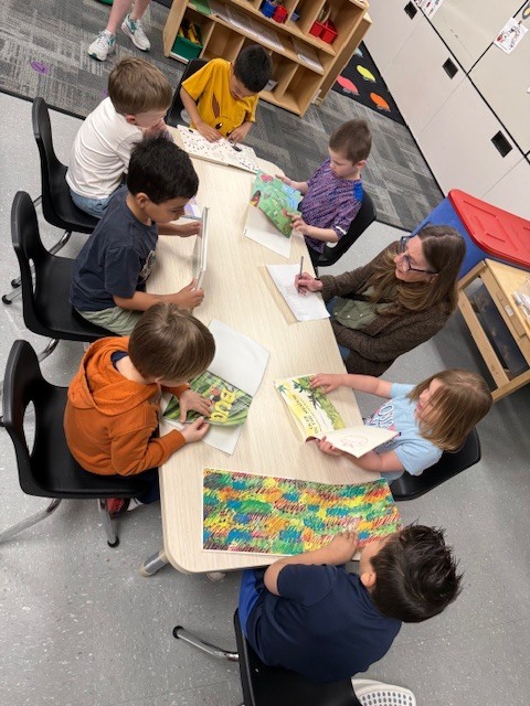 Children sit around a table with a teacher, flipping through books and pointing to different text features while engaging in group discussion.
