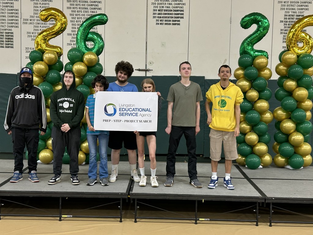 Seven people stand on a stage in front of a white wall with sports championship banners. On either side of the group are tall balloon pillars made of green and gold balloons, topped with "2" and "6" balloons.