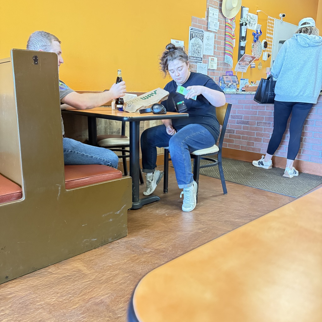 A person with dark hair in a bun wearing a black t-shirt and blue jeans sits at a restaurant table. They are looking down and holding a green smartphone over a brown paper bag. Across from them, a person in a grey shirt and blue jeans sits in a booth, holding a glass bottle. In the background, a person in a grey hoodie and black leggings stands near a counter against a brick wall. The restaurant has bright orange walls.