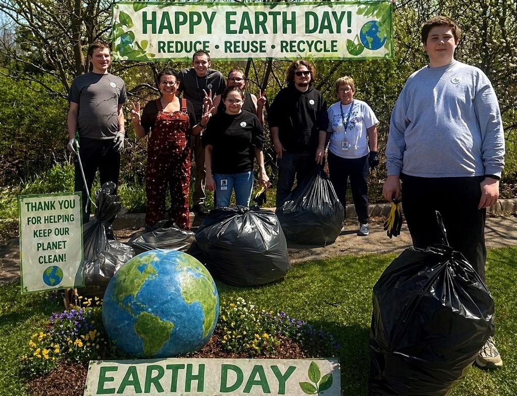 A group of eight people poses outside with full trash bags behind a large globe decoration and a "Happy Earth Day! Reduce, Reuse, Recycle" banner.
