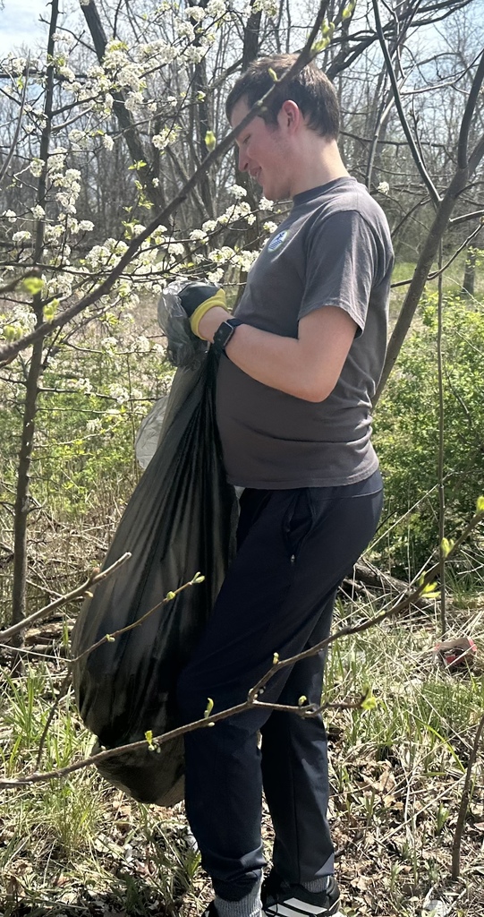 A person in a grey t-shirt and dark pants stands near a blossoming white tree, holding open a large black plastic trash bag.