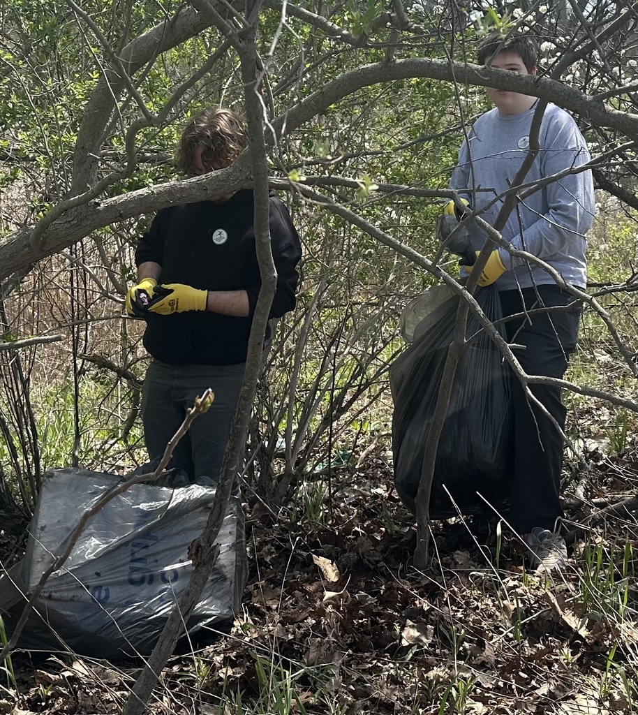 Two people wearing yellow work gloves stand among thick brush and trees, handling large trash bags during a cleanup.