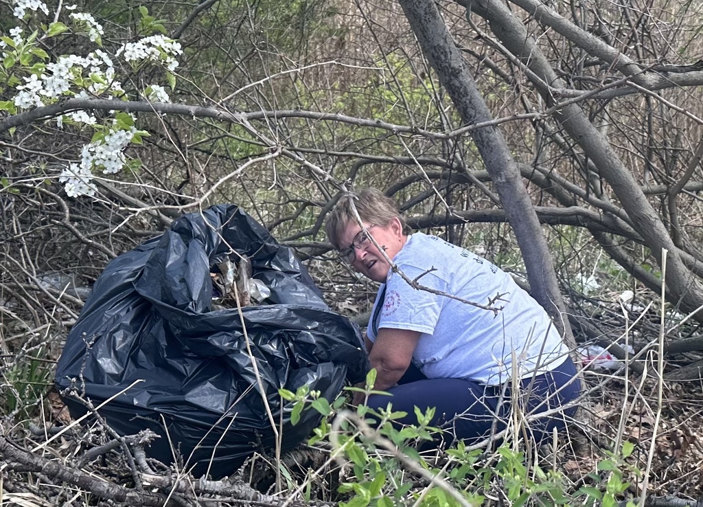 A person with glasses and a white t-shirt kneels on the forest floor next to a large, bulging black trash bag under some branches.