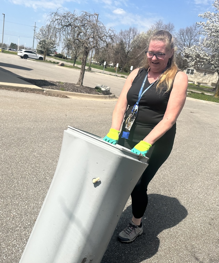 A person in a black tank top and bright green gloves tilts and moves a large, grey plastic bin in a paved parking lot