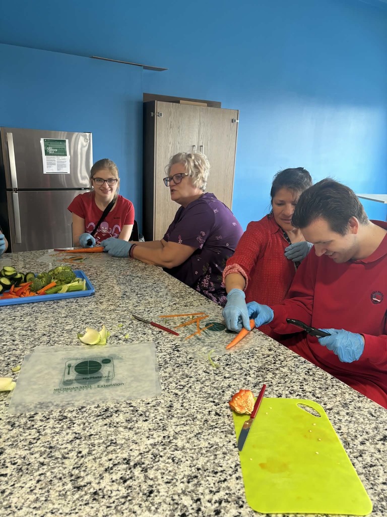 A group of people wearing blue gloves while learning to chop and peel carrots at a kitchen counter during a cooking class.