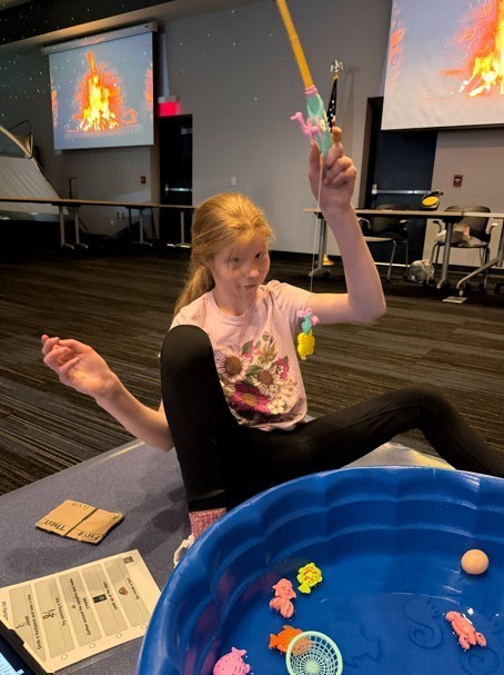 A female student sits on the floor smiling as she holds up a fishing pole. A toy fish hangs from the end of her line, which she has just "caught" from a small indoor pool filled with pretend fish.