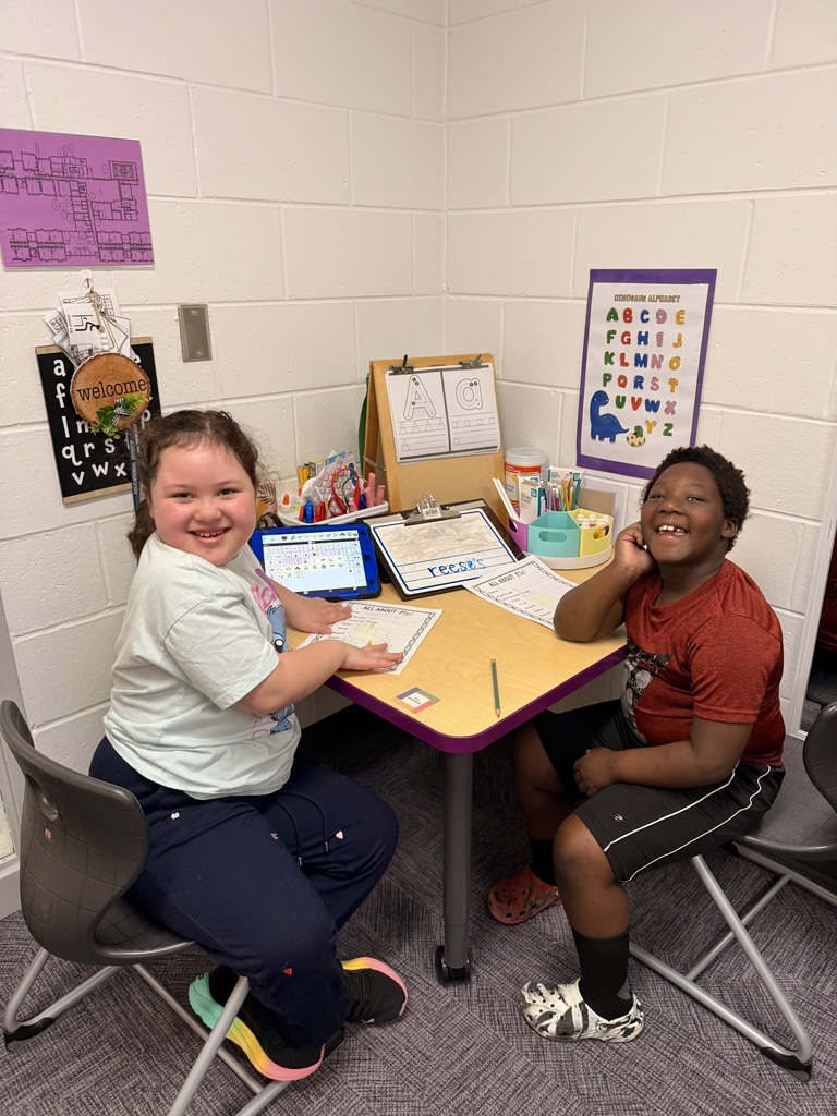 Two smiling children sit at a small, purple-edged desk in a classroom. On the left, a girl in a light blue t-shirt and dark pants rests her hand on a worksheet. On the right, a boy in a red t-shirt and black shorts smiles broadly at the camera. The desk is filled with learning tools, including a tablet displaying a communication app, a pencil carousel, and a clipboard with the word "reese's" written on it. The white cinder block walls are decorated with a floor plan and a colorful Dinosaur Alphabet poster.