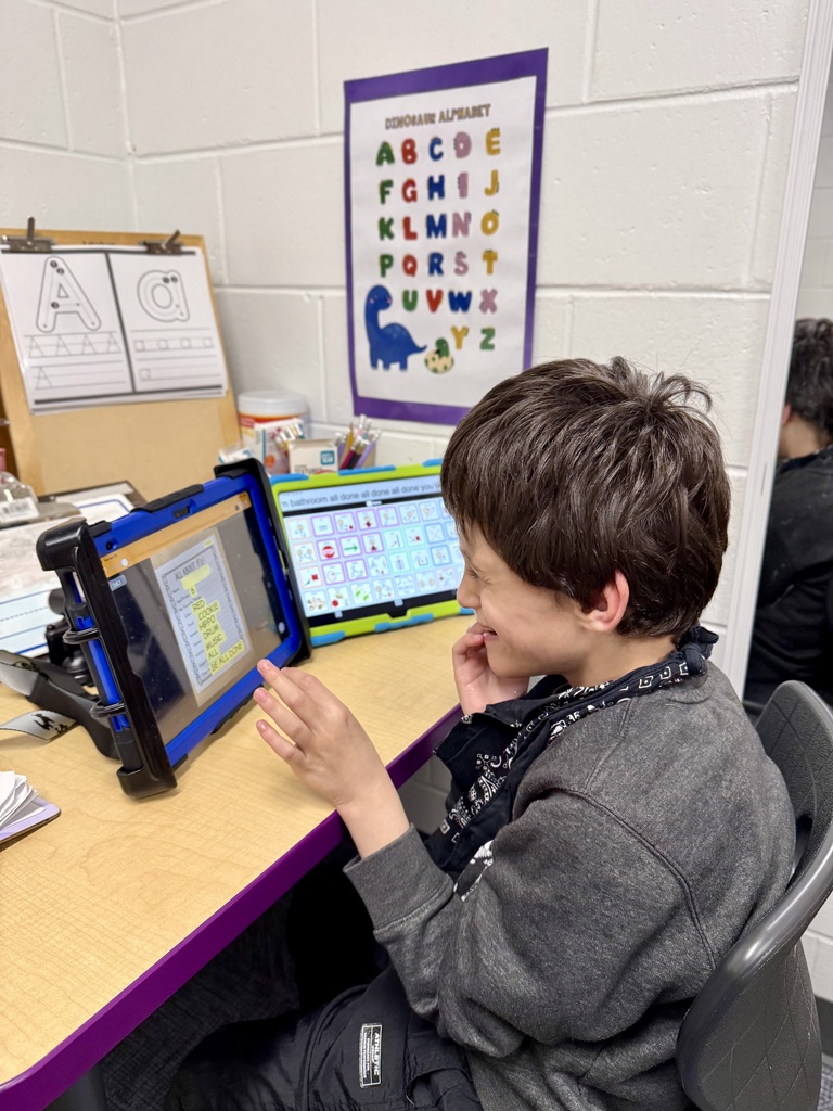 A young boy with dark hair sits at a desk, smiling as he interacts with two tablets. One tablet displays a list of simple words like "Cookie" and "Music," while the second tablet shows an AAC (Augmentative and Alternative Communication) interface with a grid of colorful icons. The classroom background features a white brick wall decorated with educational posters, including a dinosaur-themed alphabet.