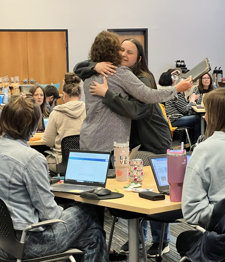 Two women embracing in a hug during a presentation in a meeting room.