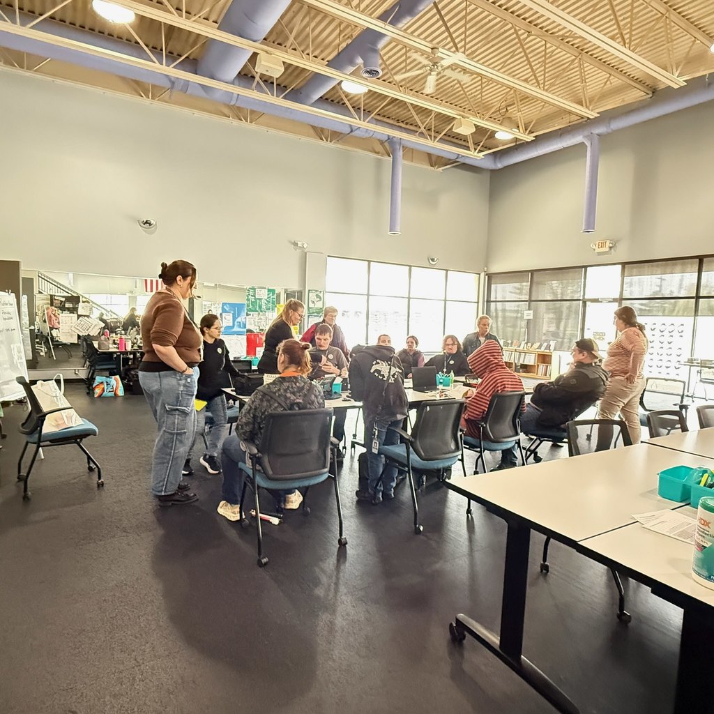 A group of people gathered around a large table in a bright, industrial-style community room or classroom, engaged in a collaborative activity or discussion.