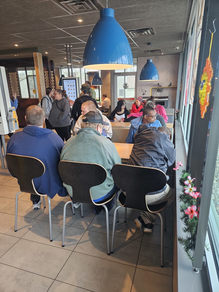 An eye-level shot indoors shows people sitting and standing at a restaurant. Three people sit on chairs in front of a long light-colored table. The person on the left is wearing a blue hoodie, the person in the middle is wearing a green jacket, and the person on the right is wearing a black jacket. More people sit on the other side of the table. To the left, several people stand in a line. In the background, there are windows and a glass door. Four blue pendant lights hang from the ceiling. A garland with pink flowers hangs on the right wall. The floor is light-colored tile.