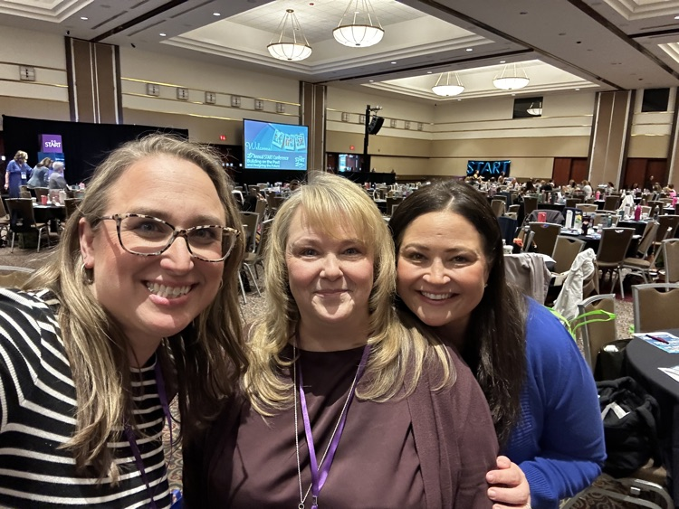 Three woman posing together and smiling in a large conference room full of people.