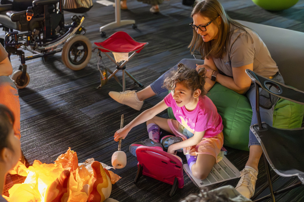 Adult helping student sit on the floor while pretending to roast marshmellows.