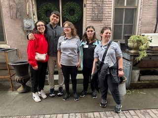 A group of five smiling young adults stands together outdoors in front of a building with a dark door and two green wreaths. They are dressed in casual clothing, including sweatshirts and polo shirts, with some wearing identification badges. The setting appears to be a courtyard or patio with brick details and potted plants.