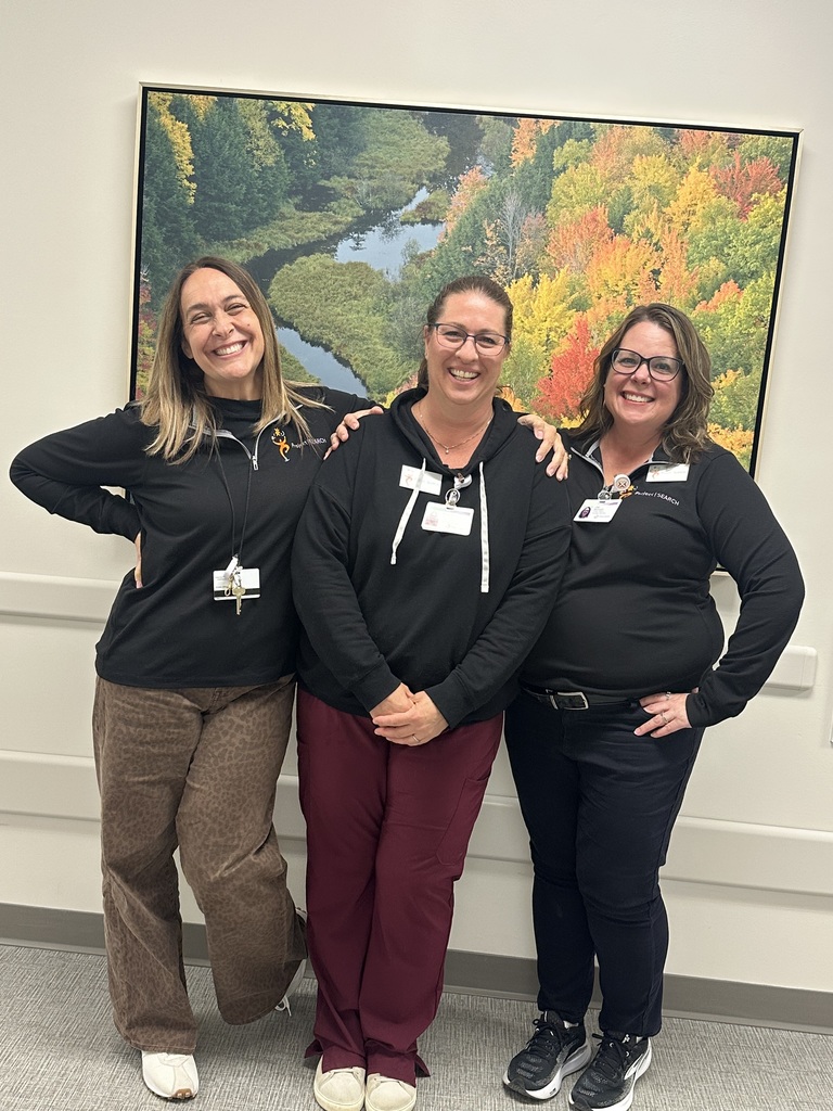 Three smiling women stand close together in front of a large framed landscape painting of an autumn forest and river. They are all wearing black tops and identification badges. The woman on the left wears leopard-print pants, the woman in the middle wears maroon scrub pants, and the woman on the right wears dark pants.