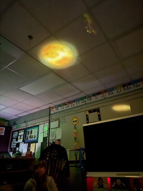 A classroom ceiling displays a projected image of the sun as preschoolers sit below and observe during a space lesson.