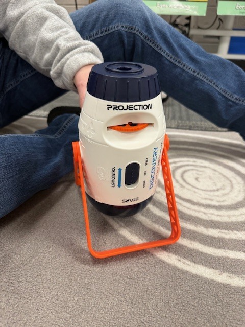 A close-up of a small classroom projector device sitting on the carpet, used to display images of planets for learning.