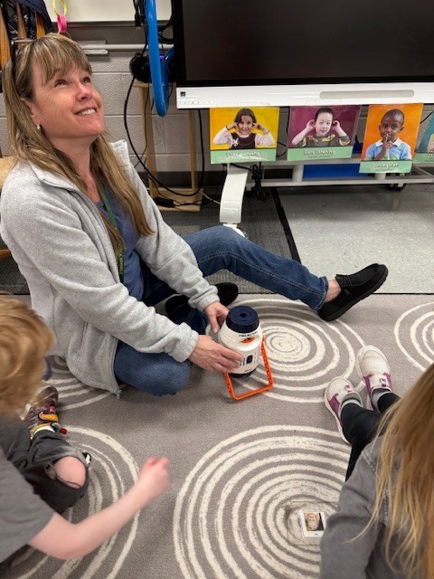 A teacher sits on the carpet with preschoolers, holding a projector as children gather around to explore images of planets together.