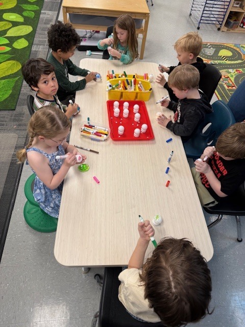 A group of preschool children sit around a table using markers and materials to decorate small round objects as they create their own planets.