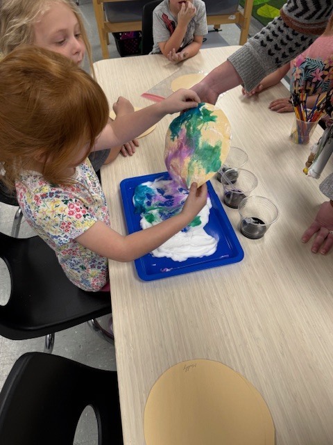 Preschool children use a paint transfer technique with shaving cream and dye to create colorful planet artwork on paper.