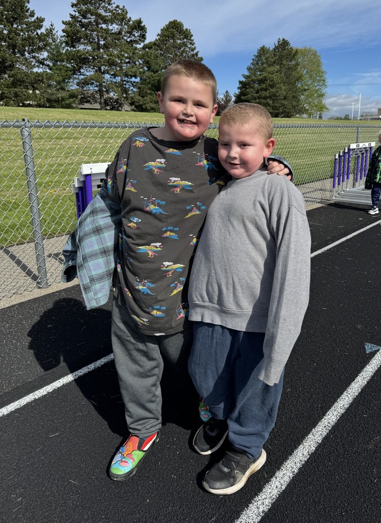 Two young boys stand together with their arms around each other on a black running track. The boy on the left wears a dinosaur-print shirt and colorful shoes, while the boy on the right wears a grey sweatshirt. They are smiling in front of a chain-link fence and a grassy field under a bright blue sky.