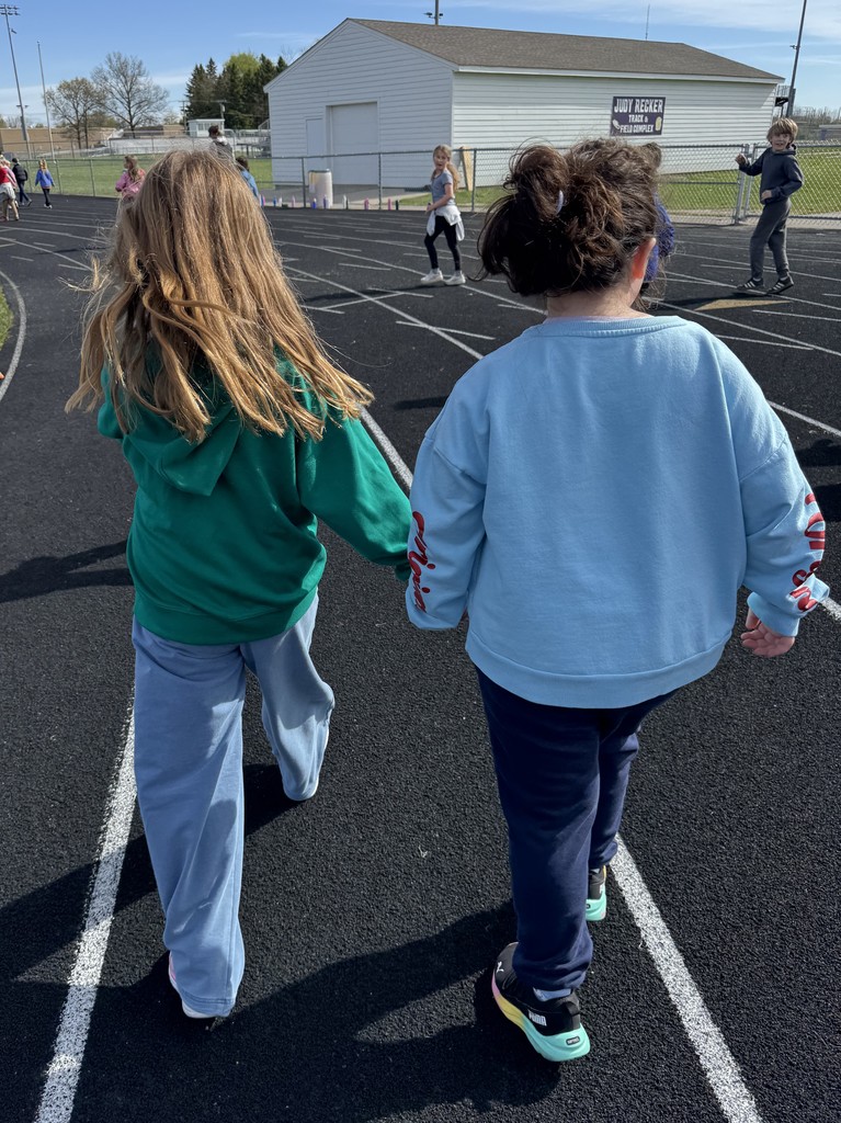 Two young girls walk away from the camera, holding hands as they stroll down a black running track. The girl on the left has long blonde hair and wears a green hoodie, while the girl on the right has dark curly hair and wears a light blue sweatshirt. In the background, other children are visible on the track near a white building.