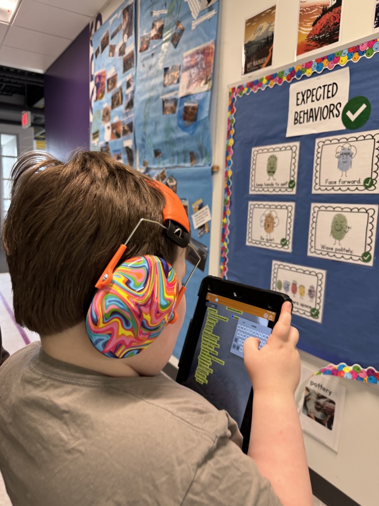 A student wearing vibrant, multi-colored swirl noise-canceling headphones types on a tablet screen in a hallway. They are standing next to an "Expected Behaviors" bulletin board that displays illustrated rules like "Keep hands to self".