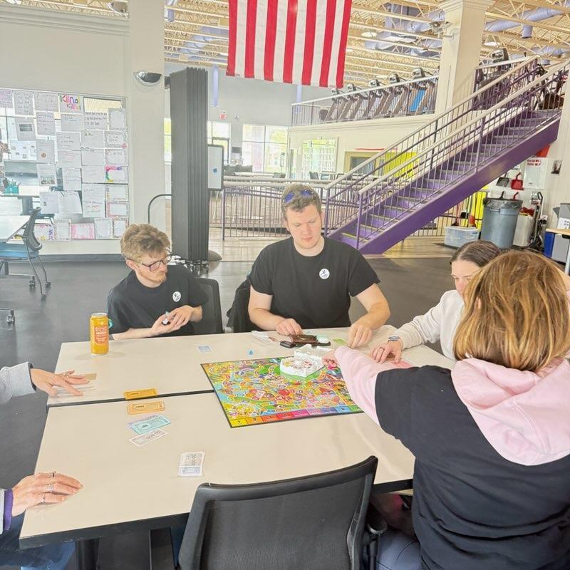 A group of young adults sitting around a white table in a brightly lit indoor space, playing the board game Life. One person holds a stack of game money while others focus on the colorful game board. In the background, there is an American flag, a purple staircase, and fitness equipment.