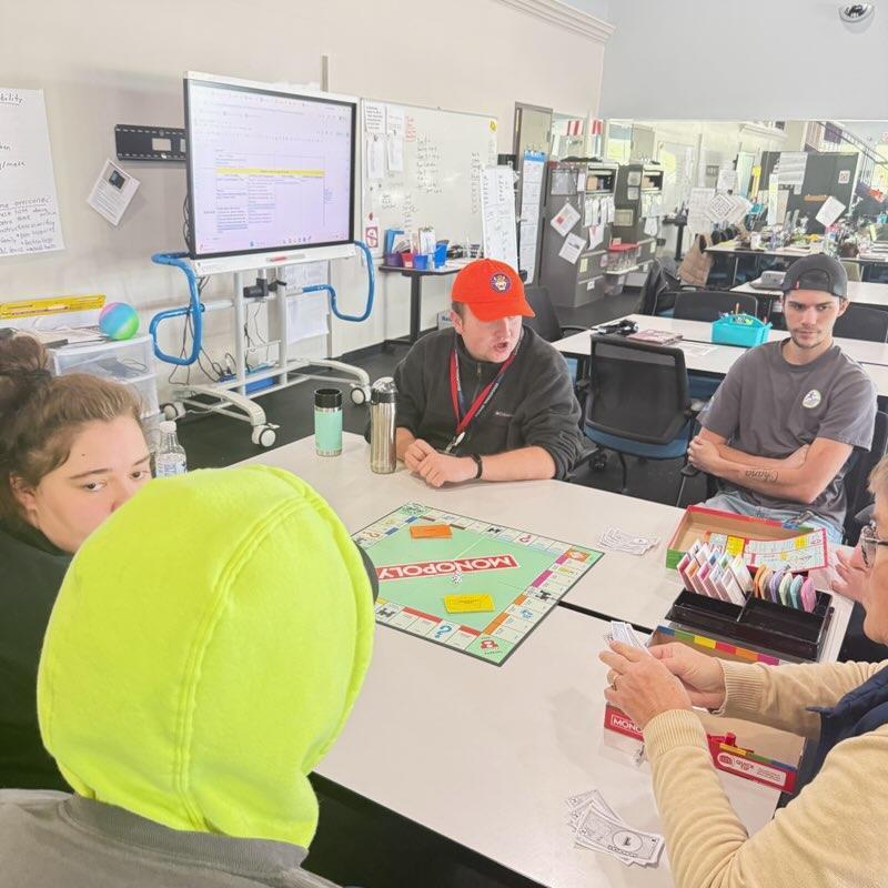 Four people are gathered around a table playing Monopoly. A young man in a bright orange hat appears to be speaking or explaining a rule, while a woman in a tan sweater manages the game's bank. A large digital screen displaying a document stands in the background of the classroom-like setting.