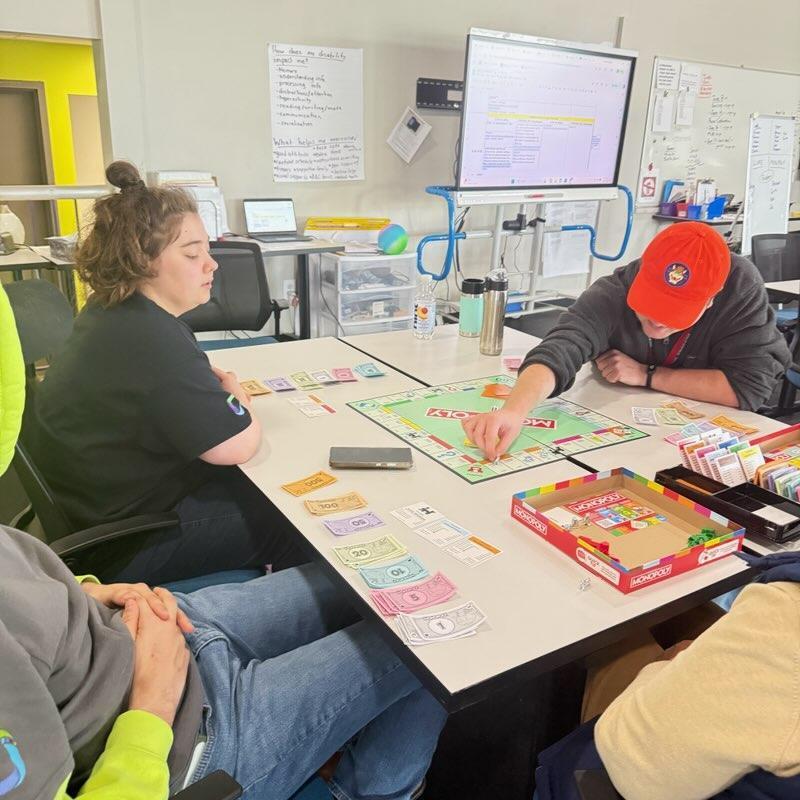 A close-up view of the same Monopoly game from a different angle. The player in the orange hat reaches out to move a game piece on the board. Neatly organized stacks of colorful Monopoly money and property cards are spread out on the table in front of the players.