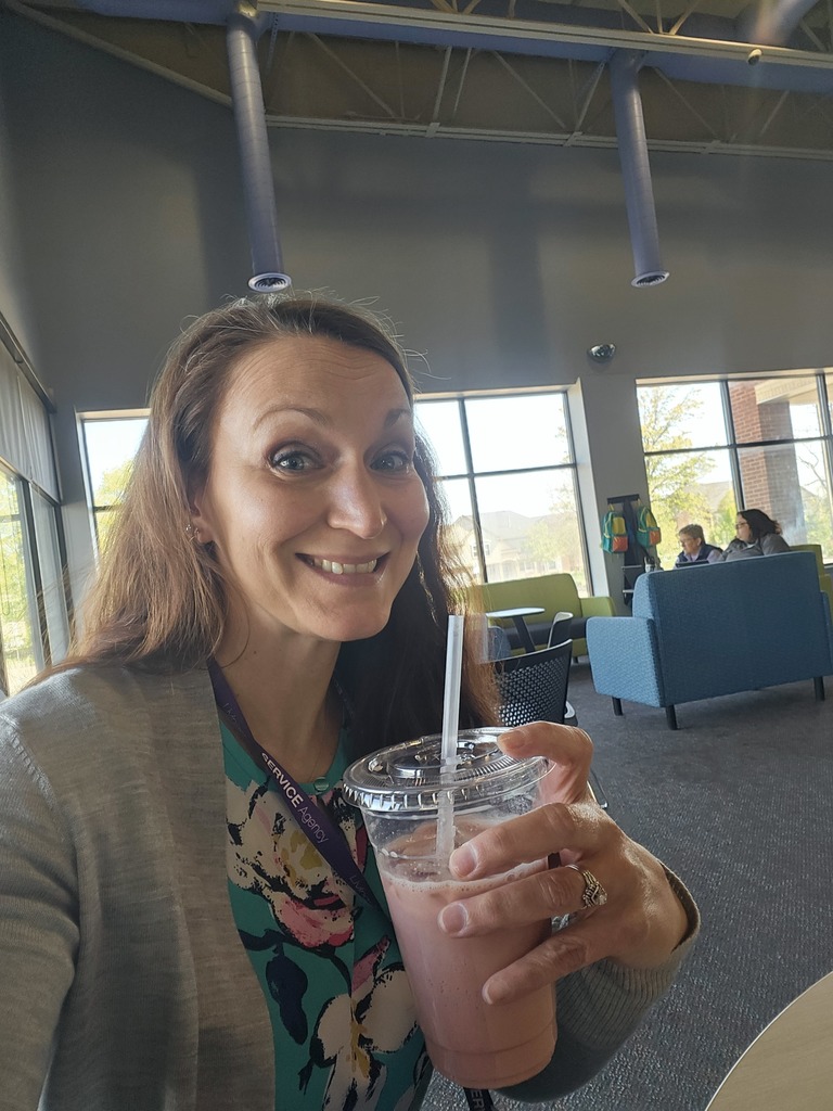 A selfie of a smiling woman with long brown hair, sitting in a bright, modern lounge area. She is holding a clear plastic cup containing a pink smoothie or blended drink with a straw.