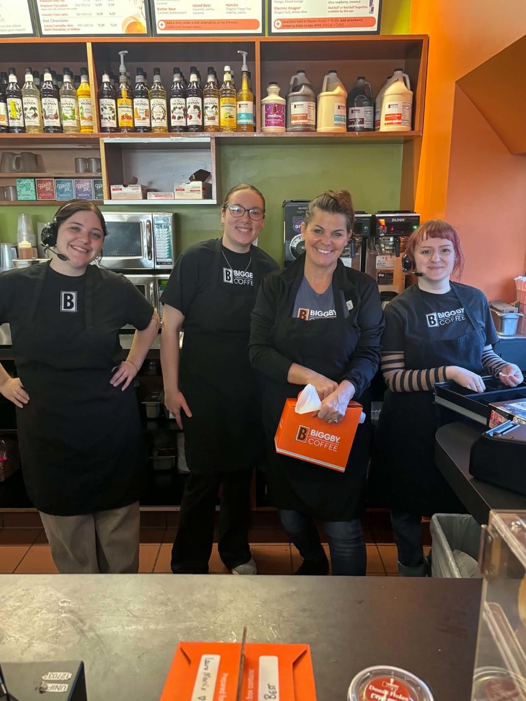 Four woman in Biggby  Coffee shirts pose together, smiling, in a coffee workplace.