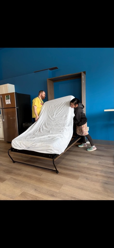 Two people work together to pull down a Murphy bed from a wall cabinet in a room with blue walls.