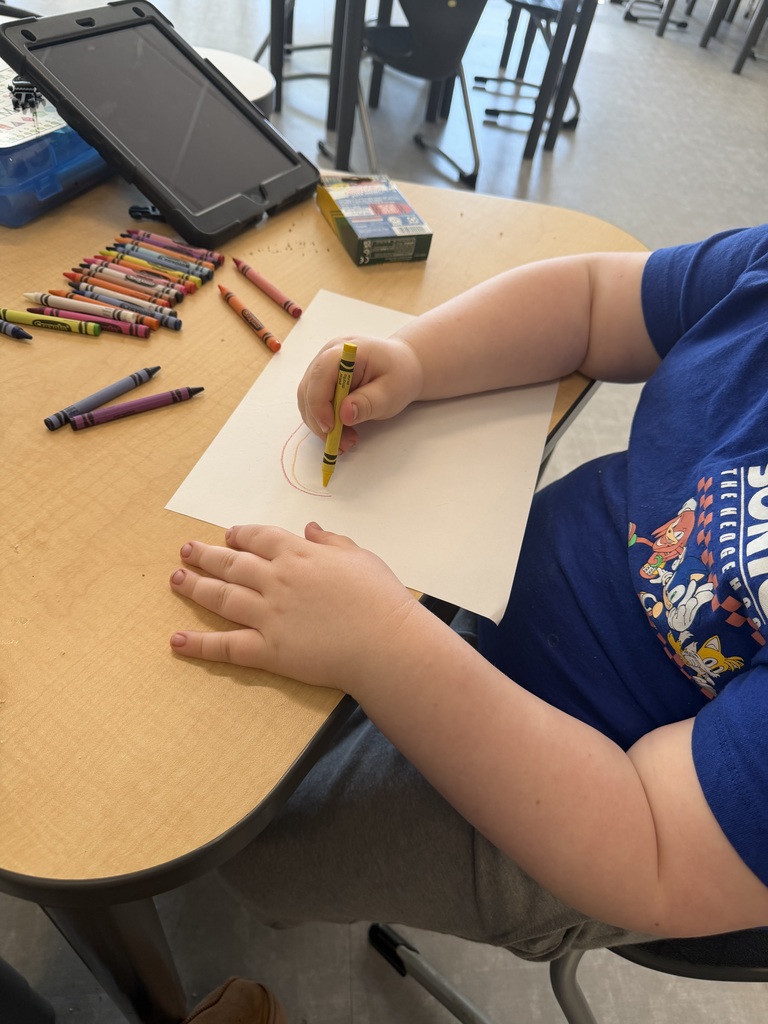 A child wearing a blue Sonic the Hedgehog t-shirt draws on a white piece of paper with a yellow crayon at a wooden table.